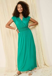 Rochie maxi verde, până la podea, dintr-un material moale, cu talie plisată, decolteu în formă de V și mâneci scurte; modelul poartă brățări suprapuse și sandale.