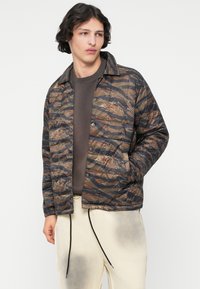 Gequilted, knoopjack in een camouflagepatroon met tinten bruin en groen. Kenmerkt zich door twee zijzakken en een ontspannen pasvorm.