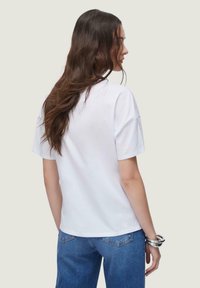 Bijela pamučna majica opuštenog kroja, kratkih rukava i zakrivljenog poruba, u kombinaciji s plavim trapericama. Glatka tekstura, minimalistički dizajn.