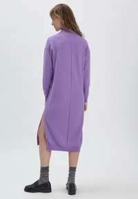 Langärmliges, lila Strickkleid mit seitlichen Schlitzen, gerippten Bündchen und lockerem Schnitt. Das Material fühlt sich weich an und hat eine glatte Textur.