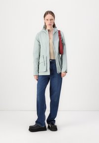 Casaco capucho verde claro, com bolsos frontais, combinado com uma blusa bege e jeans azuis folgados. Acessórios incluem uma mala cruzada vermelha. Sapatos de plataforma pretos.