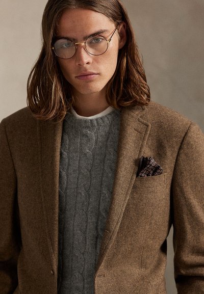 Νεαρό άτομο με καστανά μαλλιά μέχρι τους ώμους και στρογγυλά γυαλιά, φορώντας ένα καφέ σακάκι πάνω από γκρι πουλόβερ με πλέξη καλωδίου και πολύχρωμο μαντηλάκι στο τσέπη.