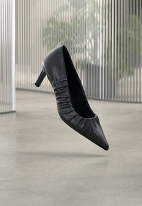 Mustast nahast kõrge kontsaga king, millel on terav nina ja ülalt voldiline tekstuur. Sile viimistlus; mõõdukas kontsakõrgus.