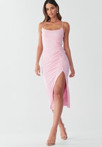 Midi-kjole i lyserød med rynkedetalje, tynde stropper og sidesprække, lavet af glat stof. Brugt med transparente hælsandaler.