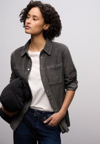 Grijs corduroy overhemd met knoopsluiting en twee borstzakken, gedragen over een wit T-shirt, gecombineerd met donkerblauwe jeans en een zwart buitenjack.