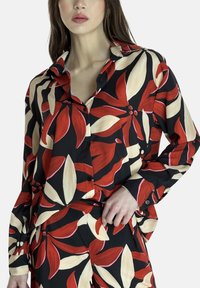 Chemise noire avec un motif floral audacieux rouge et crème, design boutonné, manches longues et coupe décontractée. Pantalons assortis visibles.