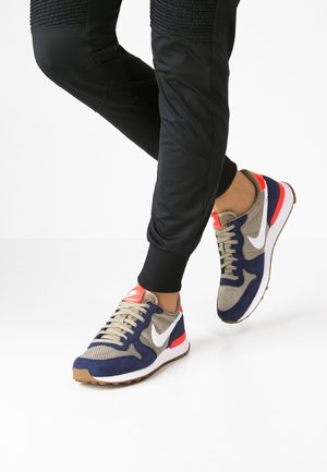 Personne portant un jogging noir et des baskets Nike multicolores avec des détails beige, marine, blanc et rouge, debout sur un sol blanc.