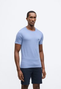 Světle modré tričko s krátkým rukávem z měkké látky, s kulatým výstřihem a malým černým logem na levé straně hrudi. Kombinováno s tmavými šortkami.