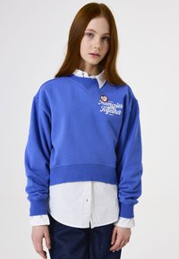 Sudadera recortada azul con dobladillo y puños acanalados, que presenta el texto blanco "Recuerdos Juntos" en el pecho, superpuesta sobre una camisa blanca con cuello.