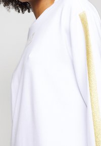 T-shirt bianco in cotone con una striscia di glitter dorato lungo la spalla e un piccolo logo sul petto. Fit morbido e casual.