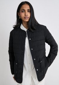 Svart kviltad jacka med rund halsringning, tryckknappar, sidofickor och dragsko-detalj. Bärs över en vit randig skjorta.