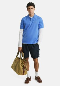 Blå pikétröja över en vit långärmad skjorta, marinblå cargo-shorts, bruna skor, håller en grön kanvas duffelväska.