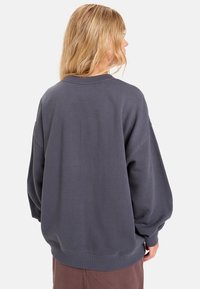 Sudadera gris de gran tamaño con cuello redondo y hombros caídos. Textura suave y puños acanalados, mostrada desde la parte de atrás.