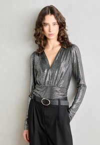 Metalni srebrni crop top s dugim rukavima i dubokim V-izrezom, s nabranim strukom, u kombinaciji s crnim hlačama visokog struka i okruglom kopčom za remen.