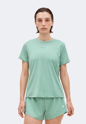 Γυναίκα με κοντά βρεγμένα μαλλιά που φοράει απλό ανοιχτοπράσινο κοντομάνικο μπλουζάκι και αθλητικό σορτς σε λευκό φόντο.