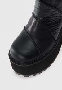 Primer plano de la punta de un zapato de plataforma negro con paneles de cuero texturizado y suave, y suela gruesa cosida, sobre un fondo blanco.