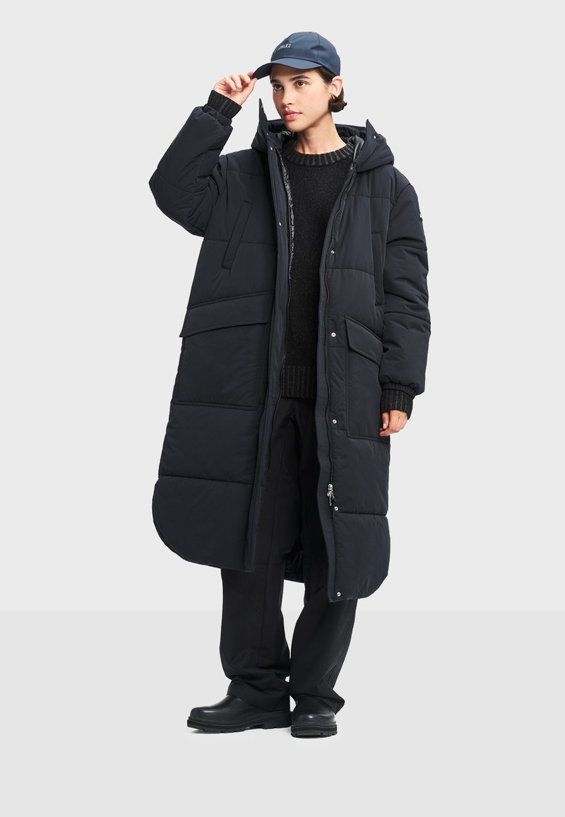 Čierny dlhý pufový kabát s kapucňou, so zipsom vpredu, viacerými veľkými vreckami a rebrovanými manžetami, nosený cez čierny sveter a nohavice.