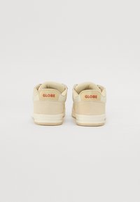Beige sneakers van suede en mesh materialen, met een laag model, een ronde neus en een rode "GLOBE" logo op de hiel.
