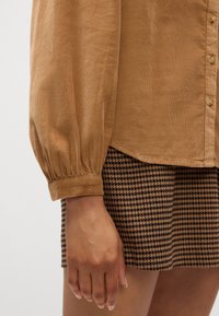 Camicia in corduroy marrone con maniche a sbuffo e chiusura a bottoni. Abbinata a una gonna a scacchi in marrone e nero.