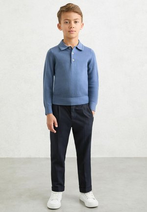 Mėlynas megztas ilgomis rankovėmis polo marškinėlis su apykakle ir sagų juosta, derinamas su siūtomis tamsiomis kelnėmis ir baltais sportbačiais.