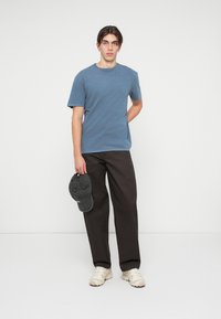 T-shirt en coton bleu à manches courtes, associé à un pantalon ample marron foncé. Le modèle tient une casquette grise et porte des baskets beiges.