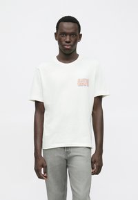 Νεαρός άνδρας που φοράει ένα λευκό T-shirt με τον τίτλο "David Bowie" τυπωμένο στο στήθος και ανοιχτούς γκρι τζιν, στέκεται μπροστά σε �απλή φόντο.