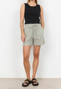 Kobieta w bezrękawniku czarnym, w jasnoszarych szortach paperbag z wysokim stanem i wiązaniem oraz w czarnych sandałach na klamrę, stojąca na szarym podłodze.