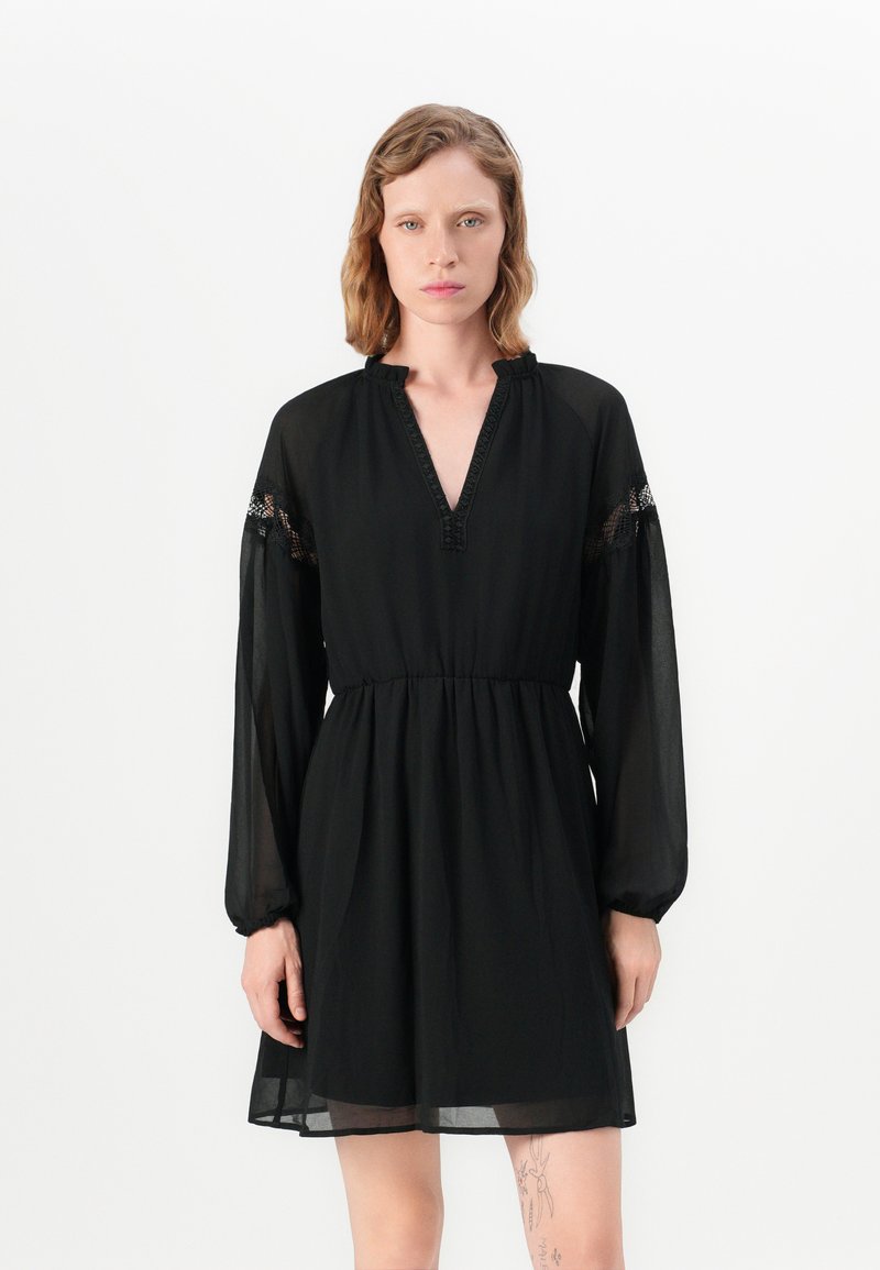 Čierne šaty vo voľnom strihu, s dlhými rukávmi, výstrihom do V a čipkovými detailmi na rukávoch. Vyrobené z ľahkej, priesvitnej látky. Pás je stiahnutý.