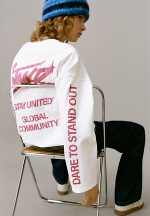 Hvid langærmet skjorte med pink tekst, med "STAY UNITED" og "DARE TO STAND OUT." Bruges med blå denim og en blåstribet hue.