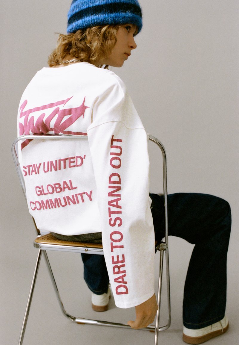 Maglietta bianca a maniche lunghe con scritte rosa, che riportano "STAY UNITED" e "DARE TO STAND OUT." Indossata con jeans blu e un berretto a righe blu.