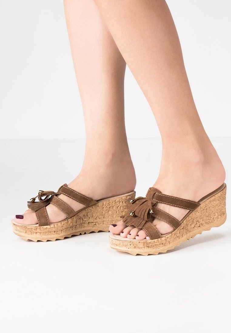 Pieds portant des sandales compensées en daim marron avec des brides croisées, des pompons et des semelles compensées à texture liège sur fond blanc.