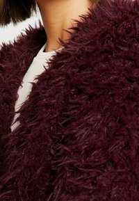Burgundský kabát z umělé kožešiny s nadýchanou texturou. Detailní záběr ukazující jemné, dlouhé vlákna a zaoblený design límce.