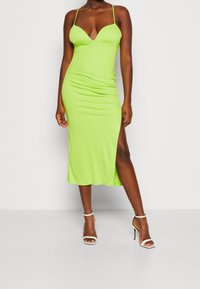 Robe midi verte moulante avec un décolleté en V profond, des bretelles fines, une fente sur le côté et des détails froncés, associée à des sandales blanches à talons hauts.