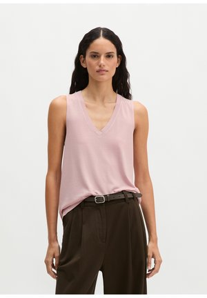 Femme aux longs cheveux foncés portant un haut sans manches rose clair à col en V, rentré dans un pantalon taille haute marron foncé avec une ceinture noire.