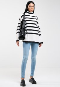 Jersey de rayas en blanco y negro con cuello de embudo, combinado con jeans skinny azul claros y zapatos negros sin cordones. Accesorio de bolso negro.