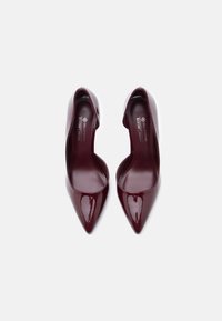 Burgundy patent leather pumps with a pointed toe, minimal design, and cutout sides. Vegan material, featuring a smooth, glossy finish.