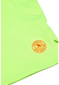 Neon žalios maudymosi šortai, pagaminti iš lengvos medžiagos, su apvaliu oranžiniu logotipu, kuriame pavaizduota banga ir tekstas. Lygus paviršius, laisvas siluetas.