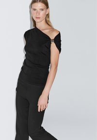 Svart, enaxlad topp med en åtsittande design. Har en rynkad ärm som betonas av ett metallklipp. Matchas med svarta åtsittande byxor.