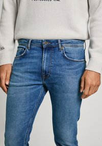 Blaue Denim-Jeans mit einer taillierten Passform, fünf Taschen und subtilen Stickdetails. Kombiniert mit einem hellgrauen strukturierten Pullover.