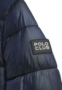 Marinblå pufferjacka med quiltade sektioner. Har en rektangulär svart lapp med "POLO CLUB" i vit text på axeln.