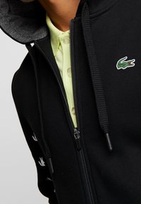 Felpa nera con zip e coulisse, con logo di coccodrillo verde, indossata sopra una camicia a polo gialla chiara.