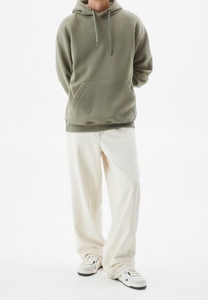 Person trägt einen übergroßen olivgrünen Hoodie, lockere weiße Hose und weiße Sneakers und steht vor einem schlichten weißen Hintergrund.