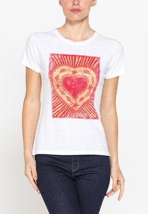 Donna che indossa una t-shirt bianca a maniche corte con motivo decorativo a forma di cuore rosso e beige sul davanti, abbinata a jeans blu scuro.