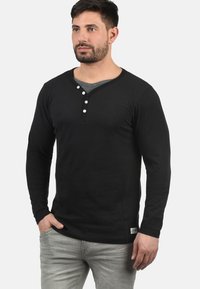 Svart langermet henley-skjorte med rund hals, tre hvite knapper på brystet, og en myk tekstur. Kombinert med lysegrå jeans.