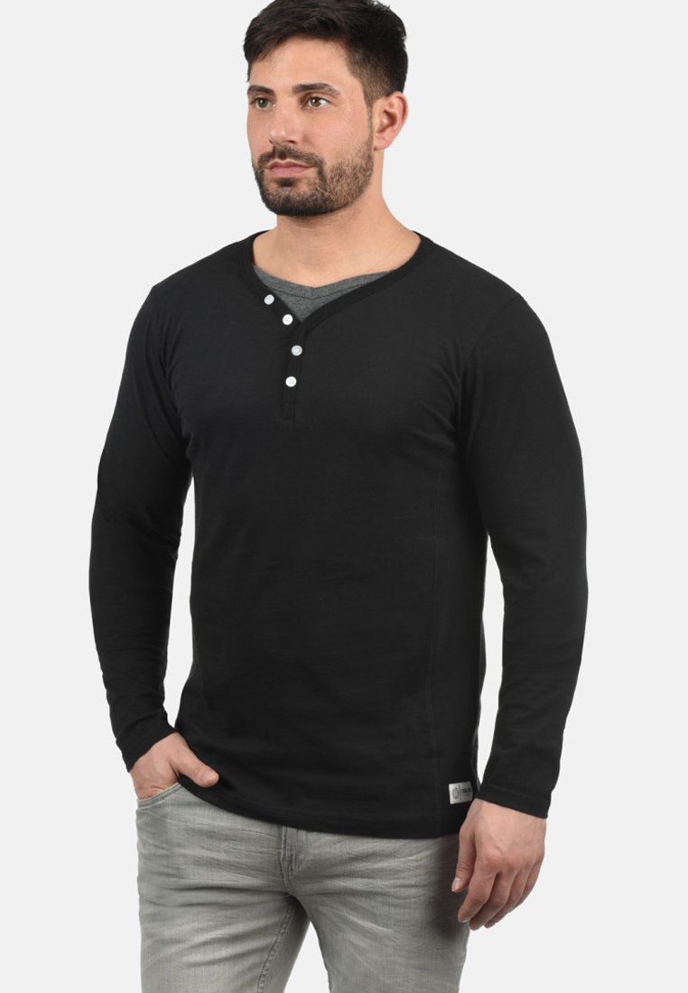 Svart langermet henley-skjorte med rund hals, tre hvite knapper på brystet, og en myk tekstur. Kombinert med lysegrå jeans.