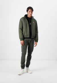 Olivenfarget bomberjakke med glidelås, ribbede mansjetter og elastisk hemming; kombinert med matchende cargo-bukser og hvite sneakers.