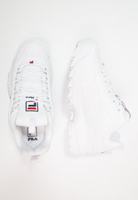 Baskets de sport blanches avec tige en cuir, design perforé, fermeture à lacets, col rembourré et semelle en caoutchouc texturée. Étiquette du logo Fila visible.