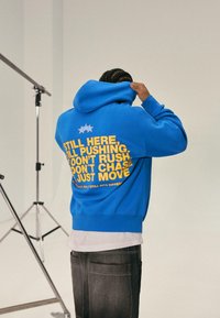Blå hoodie i bomullsmix, med gul text på ryggen: "FORTFARANDE HÄR, FORTFARANDE KÄMPANDE..." och blå stjärngrafik.