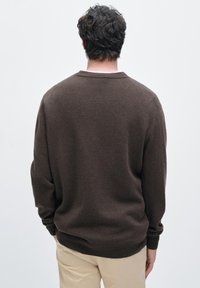 Brun stickad tröja med rund hals och långa ärmar. Mjuk textur och avslappnad passform, visad bakifrån mot en vit bakgrund.