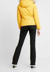 Geel, poofy jack met een capuchon en bontafwerking, gecombineerd met zwarte uitlopende jeans en witte high heels, gezien van achteren tegen een effen achtergrond.